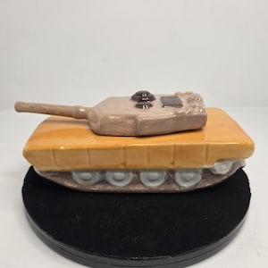 May include: A ceramic tank figurine with a tan body and a brown turret. The tank has a yellow base and gray wheels. The turret has small details and buttons. The figurine is on a black circular base.
