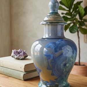 Peut inclure: Un vase en céramique bleu et or avec un couvercle. Le vase a un corps bulbeux et un long col, avec un couvercle qui a un sommet pointu. Le vase est décoré de motifs floraux bleus et or. Le vase est posé sur une surface en bois.
