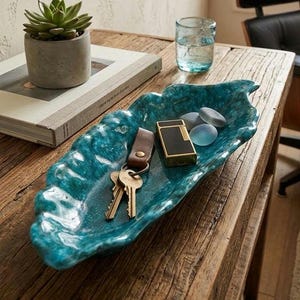 Peut inclure: Un plateau décoratif en forme de feuille, de couleur turquoise, contient des clés, un briquet et des pierres lisses. Le plateau est posé sur une table en bois rustique, à côté d'une plante succulente dans un pot en béton et d'un verre d'eau.