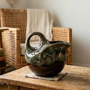 Peut inclure: Une cruche en céramique avec une base marron foncé et un dessus émaillé vert et marron. La cruche a un corps arrondi, un bec et une anse circulaire. Elle repose sur un petit dessous de verre carré sur une table en bois.
