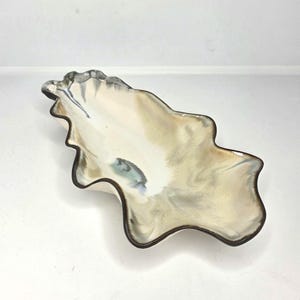 Peut inclure: Un plat en céramique allongé aux bords ondulés, présentant un intérieur crème brillant avec des traînées mélangées de brun doux, de gris et de bleu sourd. Le bord est souligné par une bordure brun foncé suivant la forme organique.