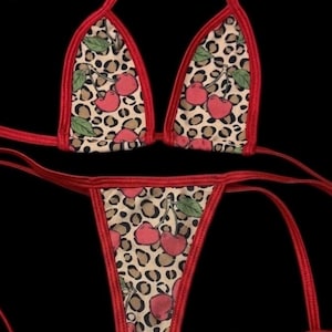 Op de afbeelding: Een tweedelige bikini met een triangel-top en een string-broekje. De stof heeft een beige achtergrond met een bruin en zwart luipaardpatroon, bedrukt met rode kersenillustraties en groene bladeren, afgewerkt met een felrode bies.
