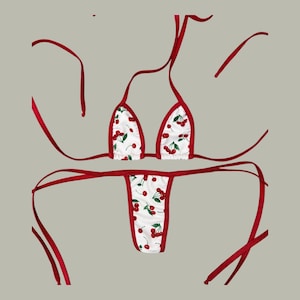 May include: A two-piece bikini set featuring a white fabric base with a repeating red cherry and green leaf print. The top consists of two triangular cups with red trim and long tie strings. The bottom is a thong style with matching red trim.