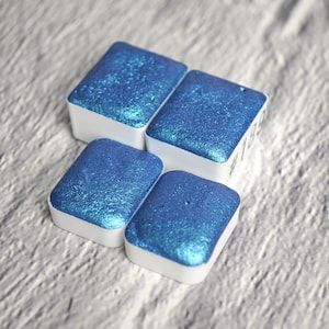 Może przedstawiać: Cztery małe kwadratowe pojemniki z farbą akwarelową o niebieskim, opalizującym połysku. Pojemniki są ułożone w kształt kwadratu.