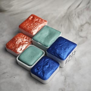 Könnte beinhalten: Fünf Aquarellfarben in weißen Plastikbehältern. Die Behälter sind in einem Halbkreis angeordnet. Die Farben sind ein metallisches Kupfer, ein blaugrünes Grün, ein metallisches Blau, ein blaugrünes Grün und ein metallisches Blau. Die metallisch blaue Farbe hat die Nummer 243.