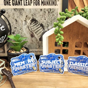 May include: Three blue and white stickers with the words "MEMORY MASTER", "SUBJECT MASTER", and "CLASSICAL MEMORIZER". The stickers have a science theme with illustrations of astronauts and planets. The background includes a globe, a house-shaped decoration, and a newspaper clipping.