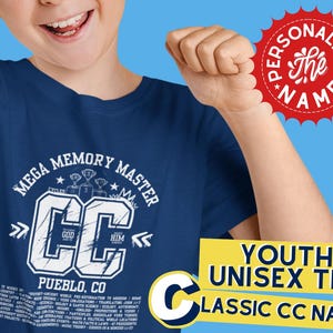 May include: A navy blue youth unisex t-shirt with a white graphic design that says "MEGA MEMORY MASTER" and "CC" with a trophy design above the letters. The text "PUEBLO, CO" is below the graphic. The shirt is shown on a child wearing it.