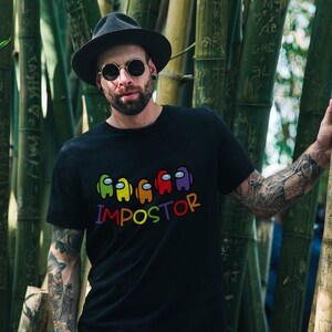May include: A person wearing a black t-shirt with a rainbow colored graphic of the characters from the game Among Us. The text "IMPOSTOR" is printed below the graphic. The person is wearing a black hat and sunglasses and has tattoos on their arms.