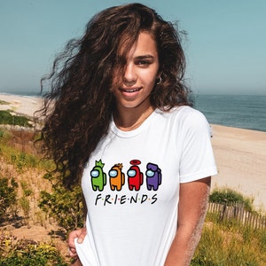 May include: White t-shirt with a graphic of four colorful Among Us characters and the word "FRIENDS" underneath. The person wearing the shirt is standing in front of a beach.