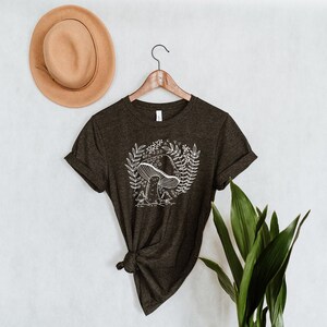 Puede incluir: Una camiseta verde oscuro con un gr&aacute;fico de hongo blanco y detalles florales. La camiseta est&aacute; en una percha y tiene las mangas enrolladas.