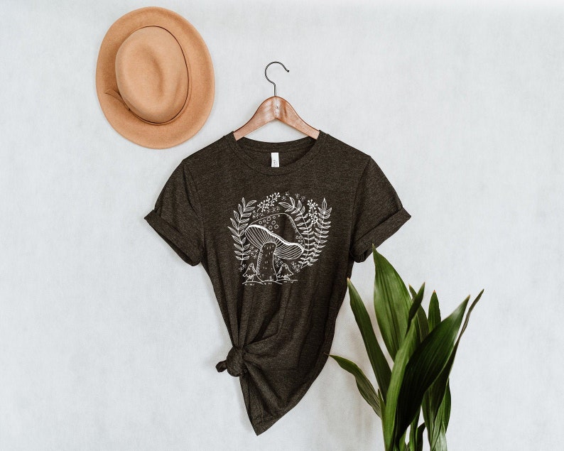 Puede incluir: Una camiseta verde oscuro con un gr&aacute;fico de hongo blanco y detalles florales. La camiseta est&aacute; en una percha y tiene las mangas enrolladas.