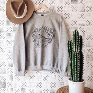 Puede incluir: Sudadera gris con una ilustraci&oacute;n en blanco y negro de un hongo con hojas y flores. La sudadera est&aacute; colgada de una percha sobre un fondo blanco con un patr&oacute;n geom&eacute;trico negro.