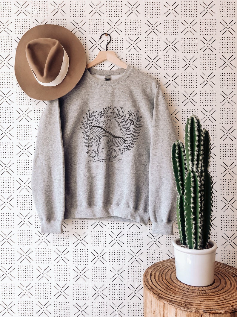 Puede incluir: Sudadera gris con una ilustraci&oacute;n en blanco y negro de un hongo con hojas y flores. La sudadera est&aacute; colgada de una percha sobre un fondo blanco con un patr&oacute;n geom&eacute;trico negro.