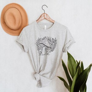 Puede incluir: Una camiseta gris jaspeada con una ilustraci&oacute;n en blanco y negro de un hongo con hojas y flores a su alrededor.