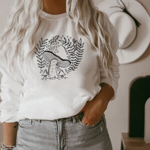 Puede incluir: Sudadera blanca con un dibujo de l&iacute;nea negra de un hongo y hojas. El texto "Shroom Queens" est&aacute; impreso en la sudadera.