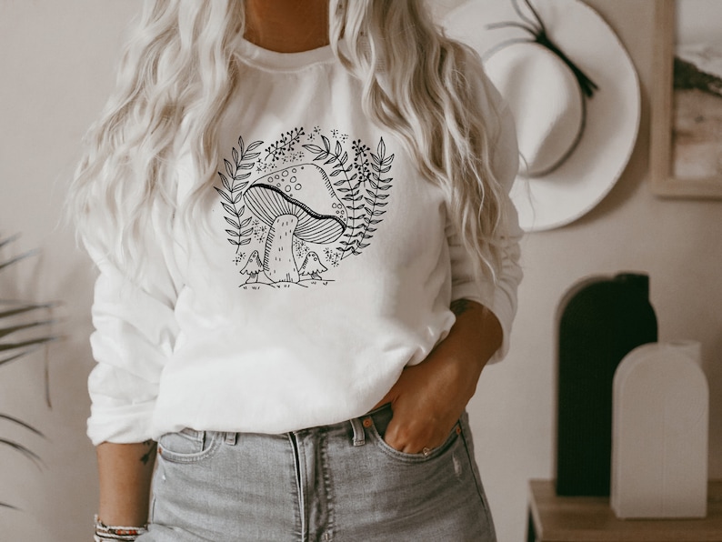 Puede incluir: Sudadera blanca con un dibujo de l&iacute;nea negra de un hongo y hojas. El texto "Shroom Queens" est&aacute; impreso en la sudadera.