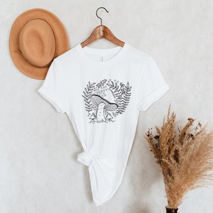 Puede incluir: Camiseta blanca con un dibujo de l&iacute;nea negra de un hongo con hojas y flores a su alrededor.