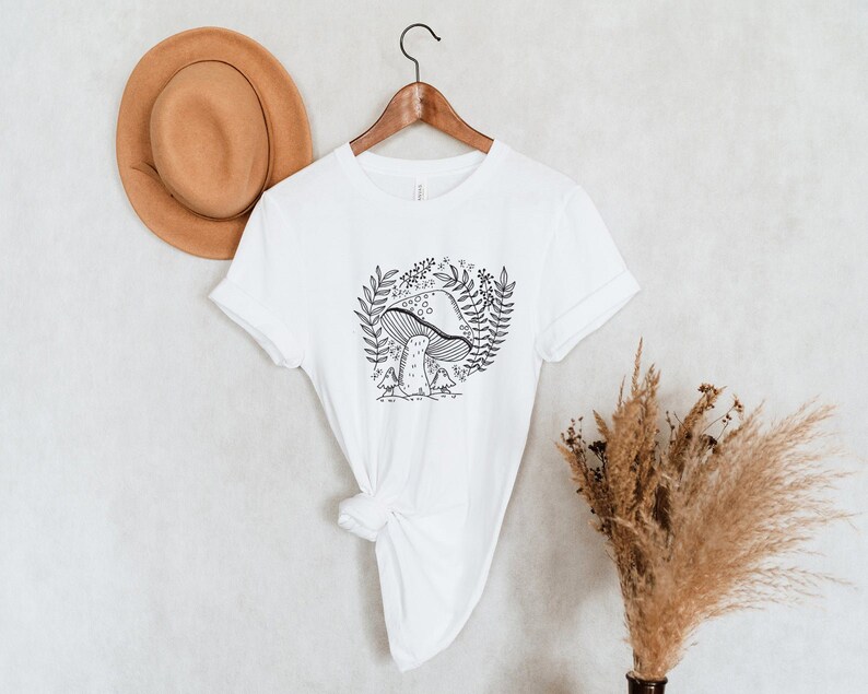Puede incluir: Camiseta blanca con un dibujo de l&iacute;nea negra de un hongo con hojas y flores a su alrededor.