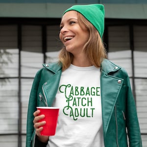 May include: A white t-shirt with green text that reads "Cabbage Patch Adult". The person wearing the shirt is smiling and holding a red cup.