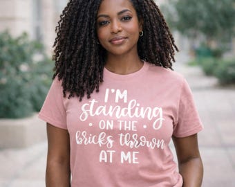 May include: A dusty rose-colored t-shirt with the text "I'M standing on the bricks thrown at me" in white script. The shirt is short-sleeved and appears to be made of a soft material.