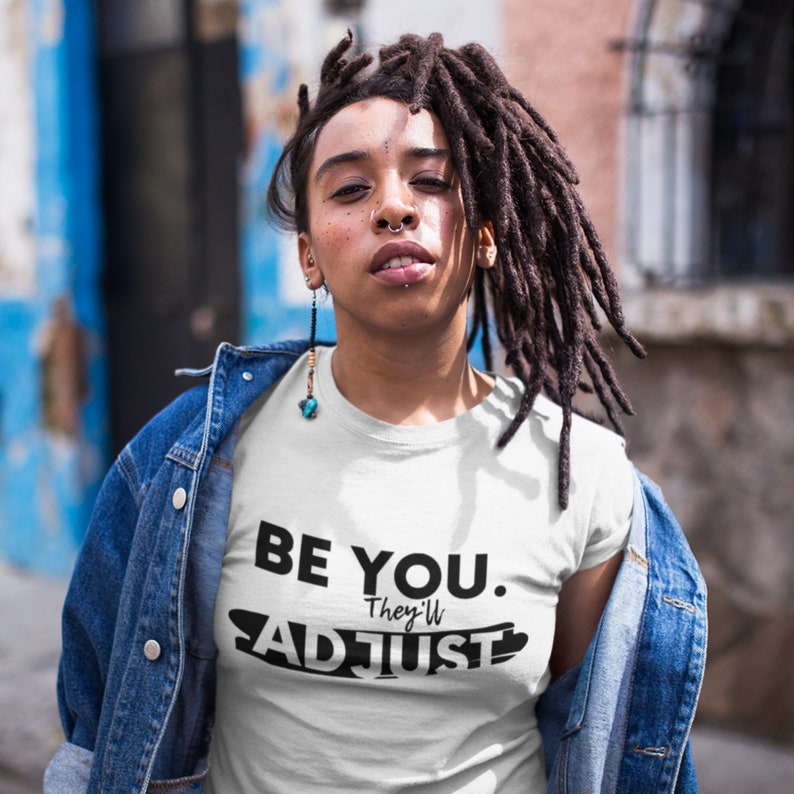 May include: White t-shirt with black text that reads "Be You. They'll Adjust". A person is wearing the t-shirt and a denim jacket.
