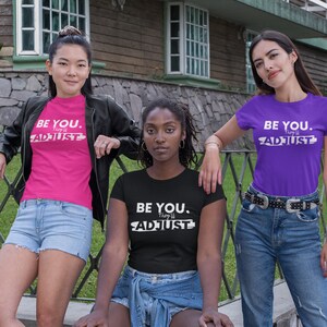 May include: Three women wearing t-shirts with the text "BE YOU. They'll ADJUST" in white letters. The shirts are pink, purple, and black. The women are standing in front of a wooden fence.