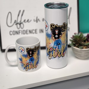 May include: A white ceramic mug and a white stainless steel tumbler with a black and yellow Pittsburgh Steelers logo and a cartoon image of a woman wearing a Steelers jersey, jeans, and a face mask. The text "Steelers Girl" is written in yellow on both mugs.