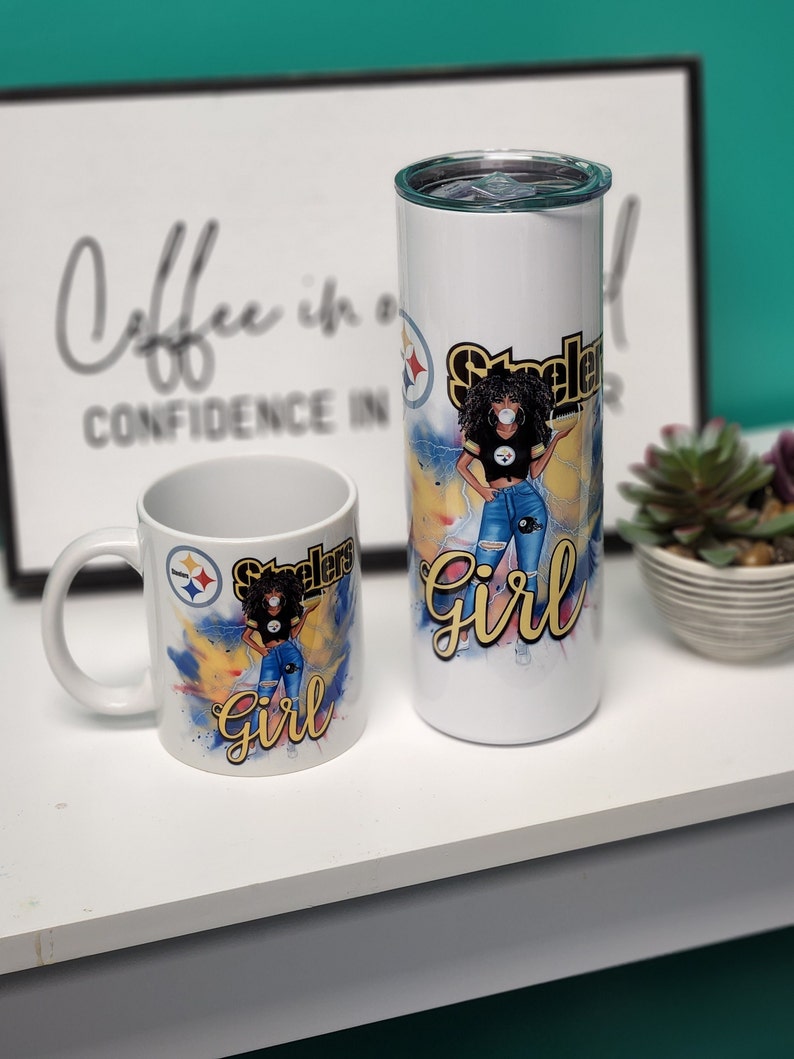 May include: A white ceramic mug and a white stainless steel tumbler with a black and yellow Pittsburgh Steelers logo and a cartoon image of a woman wearing a Steelers jersey, jeans, and a face mask. The text "Steelers Girl" is written in yellow on both mugs.