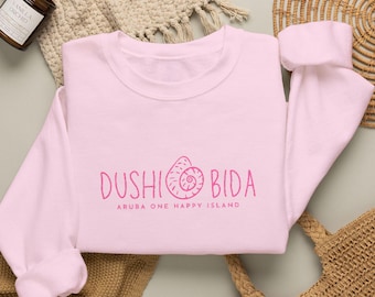 May include: A light pink crewneck sweatshirt featuring a dark pink graphic print on the chest. The text reads DUSHI BIDA with a spiral seashell icon replacing the letter O, followed by the smaller text ARUBA ONE HAPPY ISLAND below it.