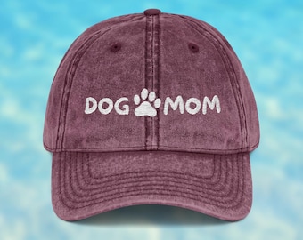 May include: A maroon baseball cap with a distressed, vintage-style finish. The front features white embroidered text that reads DOG MOM, with a white embroidered paw print icon positioned between the two words on the front panel.