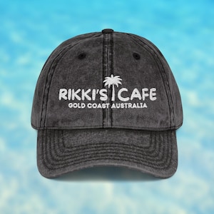 May include: A charcoal black baseball cap with a distressed finish. The front features white embroidered text that reads RIKKI'S CAFE with a palm tree icon above the word CAFE and the text GOLD COAST AUSTRALIA printed underneath in white.