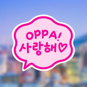 May include: A pink speech bubble sticker with the Korean phrase "OPPA! 사랑해" in bright pink lettering. The bubble has a white outline and a heart symbol. The background is a blurred cityscape.