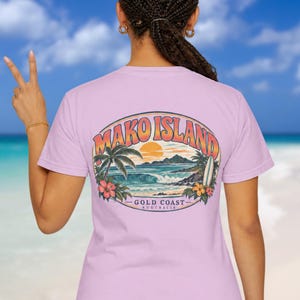May include: A light purple t-shirt featuring a back graphic of a tropical beach scene with a sunset, palm trees, a surfboard, and hibiscus flowers. The text reads MAKO ISLAND GOLD COAST AUSTRALIA in a curved, retro-style font.