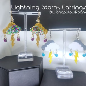 May include: A pair of lightning storm earrings with a cloud and lightning bolt design. One pair features a gold cloud with multi-colored glitter and purple raindrops. The other pair features a white cloud with gray accents and blue raindrops.  By: ShopGlowRoom