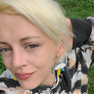 May include: A pair of earrings featuring a gray cloud with a yellow lightning bolt and blue raindrops. The earrings are worn by a person with blonde hair. The text "Classic Storm" and "By: ShopGlowRoom" is visible at the bottom of the image.