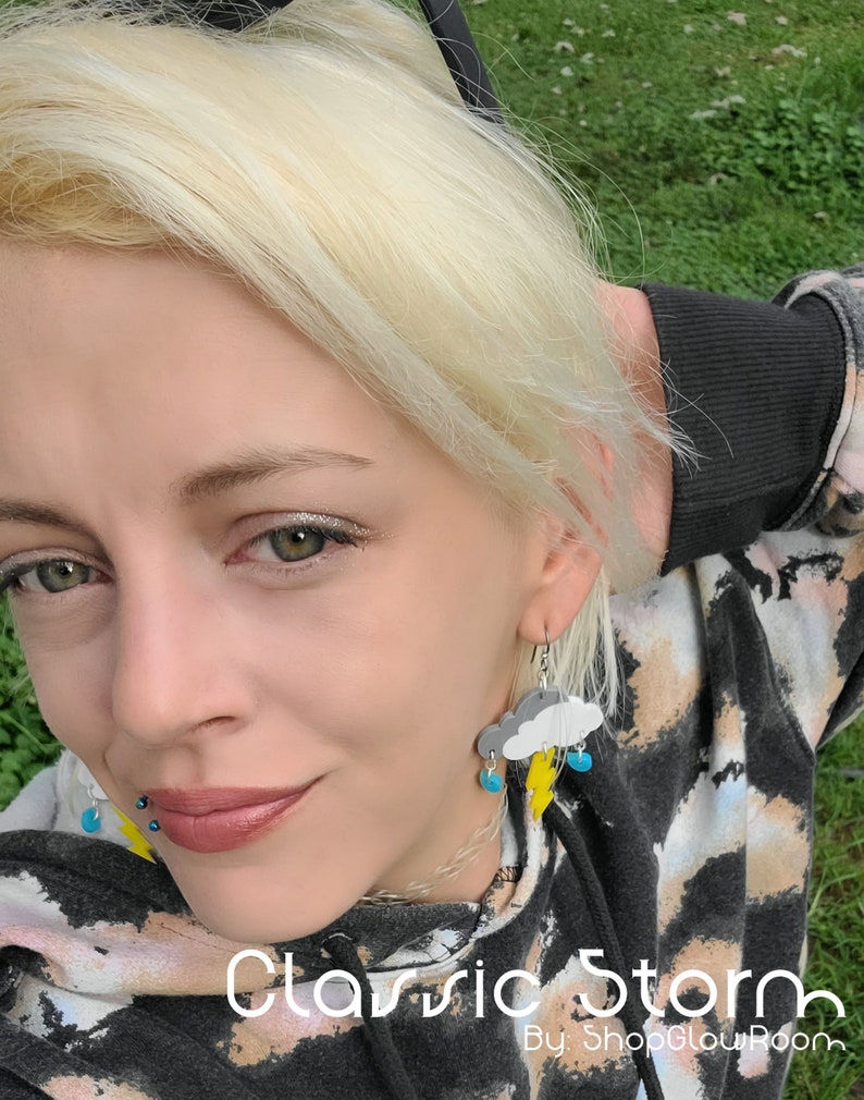 May include: A pair of earrings featuring a gray cloud with a yellow lightning bolt and blue raindrops. The earrings are worn by a person with blonde hair. The text "Classic Storm" and "By: ShopGlowRoom" is visible at the bottom of the image.