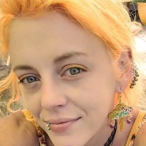 May include: A woman with bright orange hair is wearing a pair of gold and multi-colored cloud earrings. The earrings are shaped like a cloud with raindrops hanging from the bottom. The text "Purple Rain by ShopGlowRoom" is visible at the bottom of the image.
