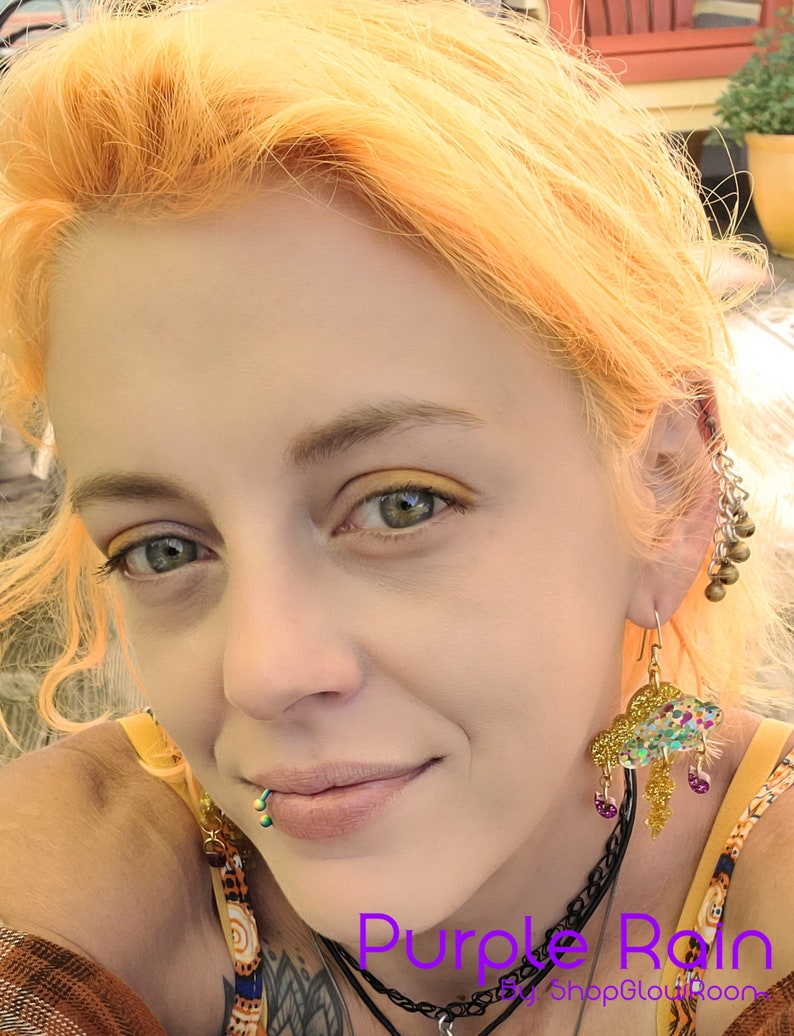 May include: A woman with bright orange hair is wearing a pair of gold and multi-colored cloud earrings. The earrings are shaped like a cloud with raindrops hanging from the bottom. The text "Purple Rain by ShopGlowRoom" is visible at the bottom of the image.