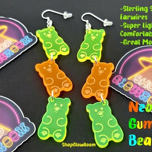 May include: Neon green, orange, and yellow gummy bear shaped earrings with sterling silver ear wires. The earrings are hanging from a white background with a neon sign that says "Shop Glow Room" and a UFO.