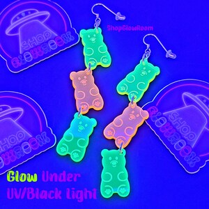 May include: Five pairs of glow-in-the-dark gummy bear earrings. The earrings are made of clear acrylic and feature a gummy bear design. The earrings are available in a variety of colors, including pink, green, yellow, and orange. The earrings are shown against a blue background. ShopGlowRoom.
