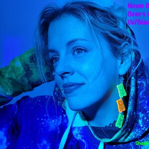 May include: A person wearing a blue and green galaxy print hoodie with neon green gummy bear earrings under a black light. The text "ShopGlowRoom" is visible in the bottom right corner of the image.