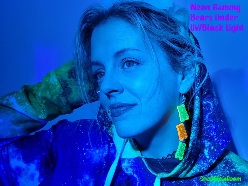 May include: A person wearing a blue and green galaxy print hoodie with neon green gummy bear earrings under a black light. The text "ShopGlowRoom" is visible in the bottom right corner of the image.