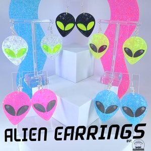 Peut inclure: Un ensemble de boucles d'oreilles en forme d'extraterrestre de différentes couleurs, dont le rose, le bleu, le vert et le noir. Les boucles d'oreilles sont en résine et contiennent des paillettes. Les boucles d'oreilles sont exposées sur un support blanc avec un fond rose et bleu.