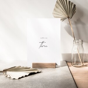 May include: A white card with the text "table no three" printed in black on a wooden stand. The card is sitting on a grey surface with a dried palm leaf on either side.