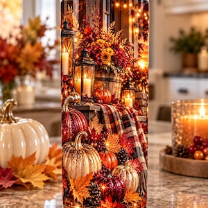 Op de afbeelding: Een cilindervormige beker met een levendige herfstprint met gloeiende lantaarns, diverse pompoenen in rood, oranje en wit, herfstbladeren, dennenappels en een plaid, allemaal verlicht door warme lichtsnoeren.