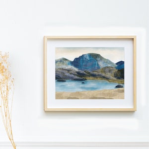 May include: A framed watercolour painting of a mountain range with a lake in the foreground. The mountains are painted in shades of blue, green, and brown, and the lake is a light blue. The painting is set against a white background.