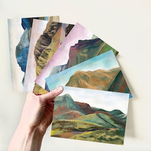 Puede incluir: Un conjunto de seis postales con pinturas abstractas de paisajes de montañas en varios tonos de azul, verde, marrón y morado.