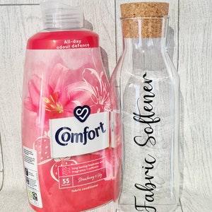 Peut inclure: Une bouteille rose et blanche d'adoucissant textile Comfort au parfum de fraise et de lys. La bouteille est étiquetée "All-day odour defence" et "55 washes". Une bouteille en verre transparent avec un bouchon en liège est étiquetée "Fabric Softener".