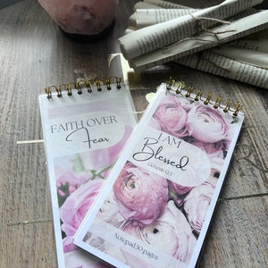 Puede incluir: Dos cubiertas de bloc de notas con diseño floral rosa y encuadernación en espiral dorada. La cubierta de la izquierda tiene el texto "FAITH OVER FEAR". La cubierta de la derecha tiene el texto "I AM BLESSED Genesis 12:3" y "Notepad 50 pages".