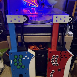 May include: Two vertical arcade controllers, one blue and one red, featuring black handles, white accents, and multiple green or white circular buttons. The blue controller has a black joystick at the bottom, while the red controller has a circular button.